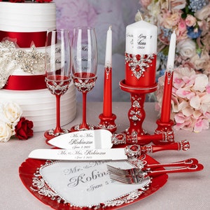 May include: Red and silver wedding cake cutting set with a heart-shaped plate, two champagne flutes, a cake knife, a fork, and two candles. The set is decorated with rhinestones and the text "Mr. & Mrs. Robinson June 5, 2023" is engraved on the cake knife and plate.