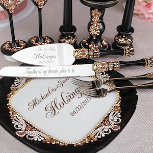 May include: A black and white cake cutting set with rose gold accents. The set includes a cake knife, a cake server, and a serving plate. The handles of the knife and server are decorated with rhinestones. The plate has a black rim and a white center with a rose gold design. The knife and server are engraved with the names "Michael & Juan Holsinger" and the date "04/22/2023".