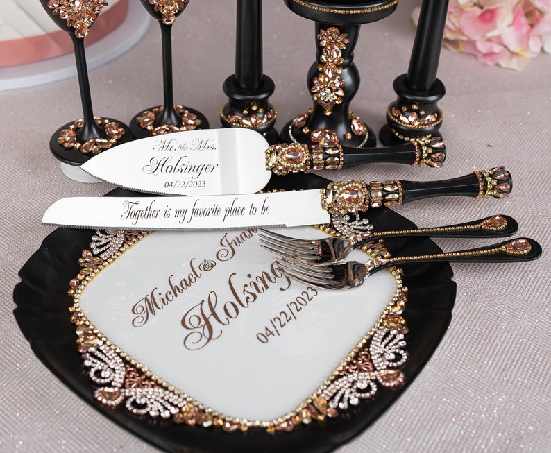 May include: A black and white cake cutting set with rose gold accents. The set includes a cake knife, a cake server, and a serving plate. The handles of the knife and server are decorated with rhinestones. The plate has a black rim and a white center with a rose gold design. The knife and server are engraved with the names "Michael & Juan Holsinger" and the date "04/22/2023".