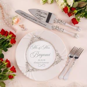May include: A silver cake knife and server set with crystal-encrusted handles, a white plate with a crystal border, and two silver forks engraved with Bride and Groom. The knife reads All that you are is all that I'll ever need and the server Mr. & Mrs. Bayomi 26/10/26.
