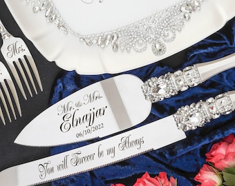 May include: A wedding cake serving set with a cake knife and server, both with crystal-studded handles. The server is engraved with "You will Forever be my Always". A white cloth with crystal trim and two forks with "Mrs." are also visible.