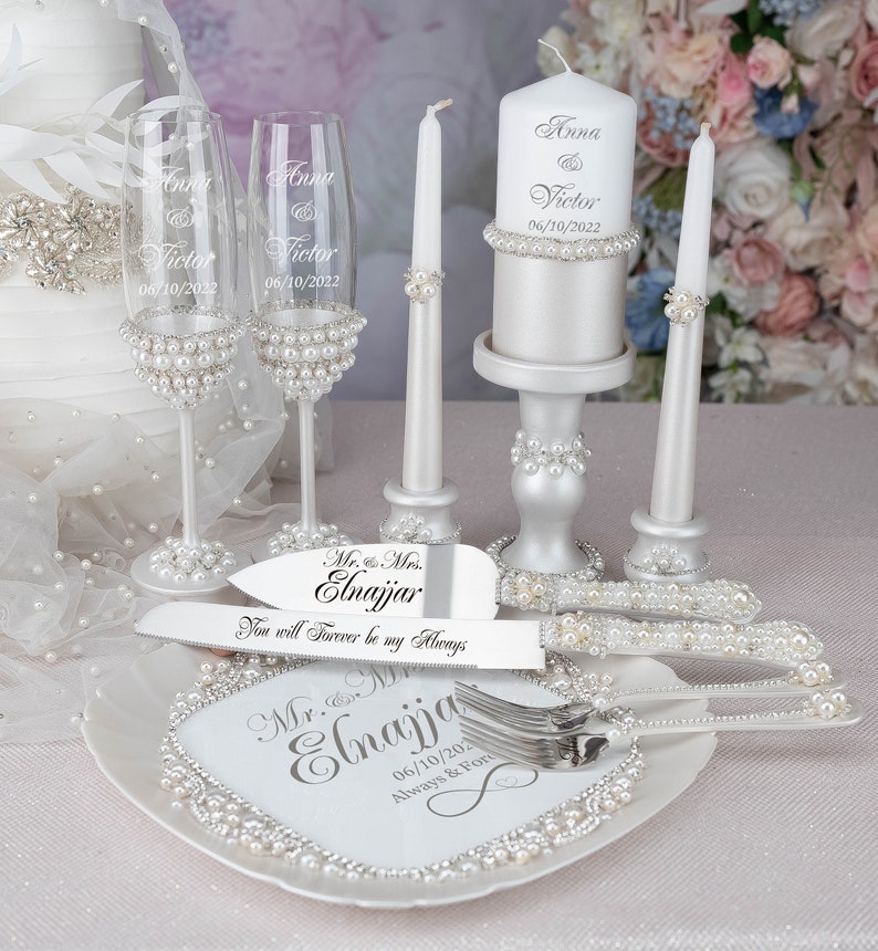 May include: Wedding cake cutting set with a white cake knife and fork, a white cake plate with pearl accents, two champagne flutes with pearl accents, and a white candle with pearl accents. The cake knife and fork are engraved with the text "Mr. & Mrs. Elnajjar You will Forever be my Always". The cake plate is engraved with the text "Mr. & Mrs. Elnajjar 06/10/202 Always & For". The champagne flutes are engraved with the text "Anna & Victor 06/10/2022". The candle is engraved with the text "Anna & Victor 06/10/2022".