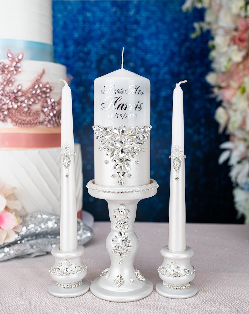 May include: A white pillar candle and two white taper candles on matching white stands. The pillar candle features the text "Mr. & Mrs. Harris 06/15/2023" and a large crystal embellishment. The stands and tapers are decorated with clear crystals.