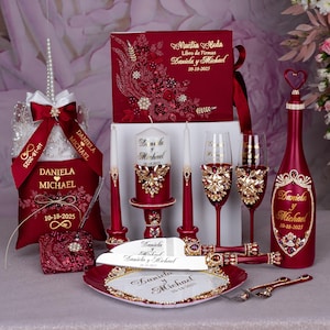 May include: A set of burgundy and gold wedding accessories, including a guest book, candles, champagne flutes, a bottle, cake server, and a ring pillow. All items are decorated with rhinestones and the names Daniela and Michael.