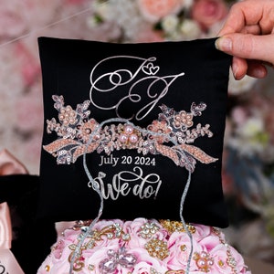 May include: Black ring pillow with a silver embroidered floral design and the text "July 20 2024 We do!" in silver. The pillow has a silver ribbon tied around it.