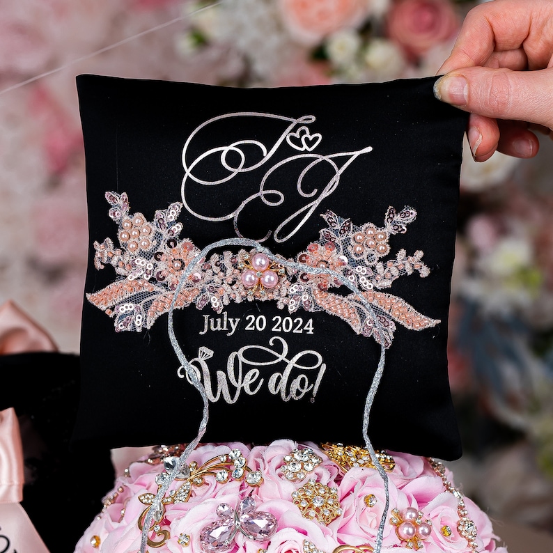 May include: Black ring pillow with a silver embroidered floral design and the text "July 20 2024 We do!" in silver. The pillow has a silver ribbon tied around it.