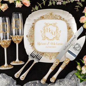May include: White cake plate with gold trim and rhinestones, featuring a gold monogram "B&M" and the text "Brian & Melanie 21/10/2023". Two champagne flutes with gold trim and rhinestones, and a cake knife and server with gold trim and rhinestones. The knife has the text "Always & Forever" engraved on it.