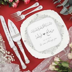 May include: A white ceramic cake plate with a silver trim and a silver rhinestone border. The plate has Arabic calligraphy and the text "Ramah Nabila 08/19/2024 It was Always you" in gray. The plate is surrounded by white flowers and greenery.