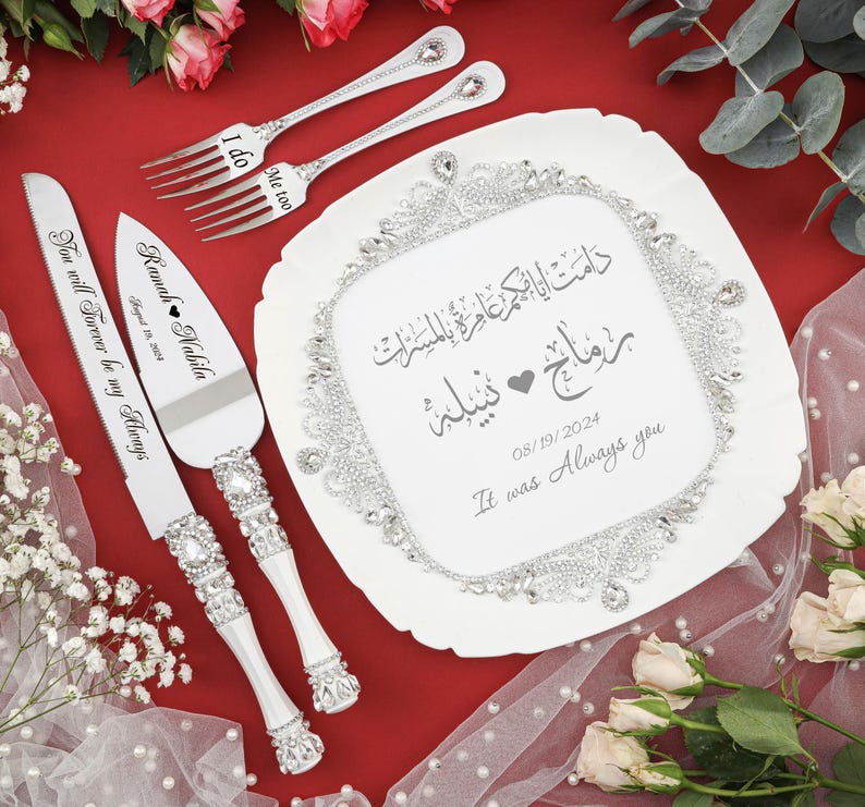 May include: A white ceramic cake plate with a silver trim and a silver rhinestone border. The plate has Arabic calligraphy and the text "Ramah Nabila 08/19/2024 It was Always you" in gray. The plate is surrounded by white flowers and greenery.