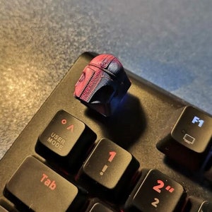Puede incluir: Un teclado negro con una tecla Mandalorian roja y azul. La tecla es una réplica impresa en 3D del casco del Mandaloriano. El teclado tiene una retroiluminación roja.