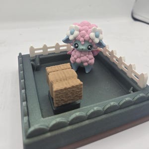 May include: A small pink and blue sheep figurine with a white flower crown stands in a miniature fenced-in area with a brown hay bale.