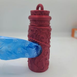 May include: A red plastic dog waste bag dispenser with an ornate design. A blue bag with paw prints is partially pulled out of the dispenser.