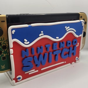 May include: A red, white, and blue Nintendo Switch stand with the words "Nintendo Switch" in blue letters. The stand is designed to hold a gold Nintendo Switch console.