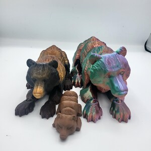 May include: Three 3D printed bear figurines. Two adult bears are sitting, one is brown and black, the other is iridescent green and purple. A smaller brown bear cub is lying down.