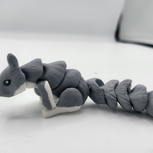 May include: A gray and white articulated squirrel figurine with a segmented tail. The figurine is made of plastic and has a textured surface.