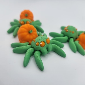 May include: Four green 3D printed spiders with orange pumpkin accents. The spiders have eight legs and large, round eyes.