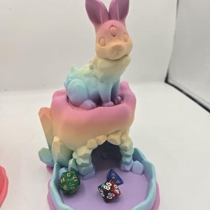 May include: A multi-colored dice tower with a bunny figure on top. The tower is in pastel colors and has a dice tray at the bottom. Several dice are in the tray. The bunny has three eyes.