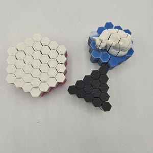 May include: Three sets of hexagonal tiles. One stack is white with red edges, another is black, and the third is blue with a white cat figurine on top. The tiles are arranged on a white surface.