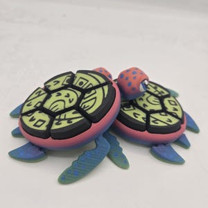 May include: Two small, colorful, 3D printed turtle figurines. The turtles are pink, green, blue, and black with a geometric pattern on their shells.