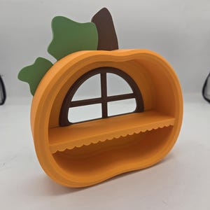 May include: A pumpkin-shaped shelf with a bright orange exterior, a brown stem, and two green leaves. The interior features a dark brown arched window frame and a horizontal shelf with a scalloped edge, providing a small display area.