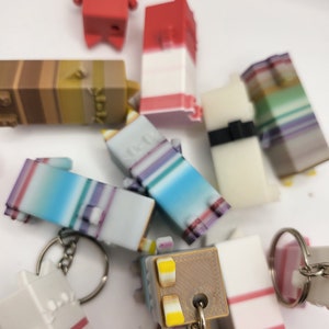 May include: A collection of colorful, layered, 3D printed keychains. The keychains are shaped like small blocks and feature various color combinations, including red, white, blue, brown, and green.