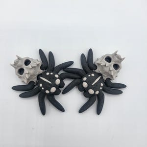 May include: A pair of black and white spider earrings with a skull detail. The spiders have a white body with black dots and a white stripe down the center. The skull is white with black eye sockets.