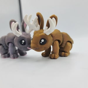 May include: Two small, brown and gray, articulated rabbit figurines with antlers. The rabbits are facing each other and appear to be touching noses.
