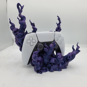 May include: A white video game controller sits on a purple 3D printed stand shaped like a skull and flames.