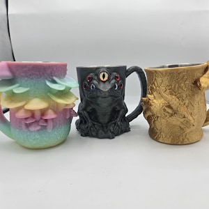 May include: Three decorative mugs with unique designs. One mug is pastel-colored with a layered, mushroom-like design. Another is black with a creature face and multiple eyes. The third is gold-colored with a dragon-like design.