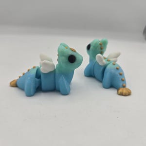May include: Two small, blue dragon figurines with white wings and gold accents. The dragons have a gradient of light blue to teal on their heads and backs. They are lying down on a white surface.