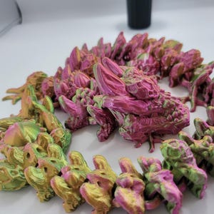 May include: A pink and green iridescent dragon figurine made of plastic. The dragon is curled up in a circle with its head resting on its tail. The dragon is made up of many small, individual pieces that are connected together.