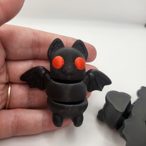 May include: A black 3D-printed bat figurine with orange eyes and small wings. The bat is held in a hand against a white background. The bat's body is segmented, and it has small feet.