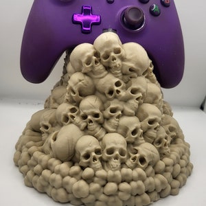 May include: A purple video game controller sits on a stand made of a pile of small, beige skulls.