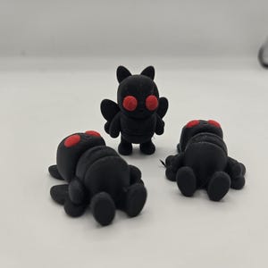 May include: A group of black figurines with red eyes. One figurine stands upright, while two others are lying down. The figurines have a cartoonish design, with bat-like wings and a rounded body. The background is plain white.