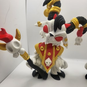 May include: A white and red 3D printed figurine of a goat-like creature with black horns and a gold staff. The creature is wearing a red and gold robe with a skull design.