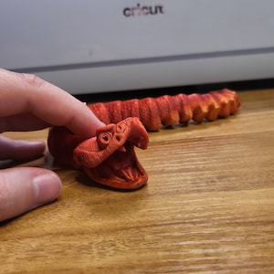 May include: A detailed, red 3D-printed snake figurine with an open mouth, held by a hand. The snake is segmented and has a textured surface. The background includes a wooden surface and a white Cricut machine.