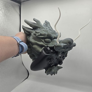 May include: A gray dragon head sculpture with a controller integrated into its design. The dragon has detailed scales and two white, curved horns. The controller is black with buttons and joysticks.