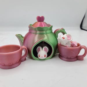 May include: A pink and green teapot with a heart-shaped opening, two pink teacups, and two white mice figurines. The teapot has a heart-shaped lid and a green and pink iridescent finish.