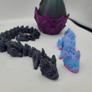 May include: Three 3D printed figurines of dragons. One is a dark gray dragon curled up in a coil. Another is a light blue and pink dragon with a light blue belly. The third is a purple egg with a dark gray dragon inside.
