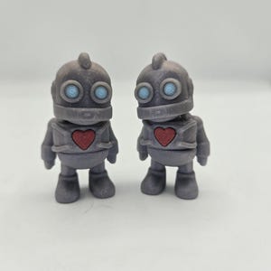 May include: Two gray robot figurines with red heart-shaped chests and light blue eyes. The robots have a classic design with rounded features and a small antenna on top of their heads. They are standing on a white surface.