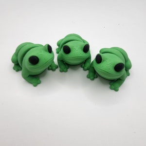 May include: Three green frog figurines with black eyes. The frogs are sitting on a white surface.