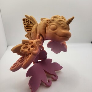 May include: A 3D-printed fantasy creature with a fish-like body, insect-like wings, and a unicorn horn. The creature is primarily tan with hints of pink and is suspended above purple leaf-shaped elements.