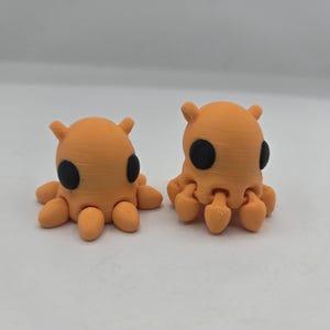 May include: Two orange octopus figurines with black eyes and rounded tentacles. The figurines have a textured surface and small, ear-like protrusions on top of their heads. They are displayed on a white surface.