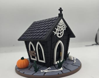 May include: A miniature black chapel with a cross on top and a white spiderweb design. The chapel has arched windows and a door. An orange pumpkin, bones, and a small white dog are on the base. The roof is dark blue.