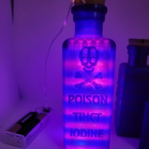 May include: A blue glass bottle with a cork stopper and a skull and crossbones design. The bottle is lit from within with pink and blue lights and has the words "POISON TINCT IODINE" printed on it.