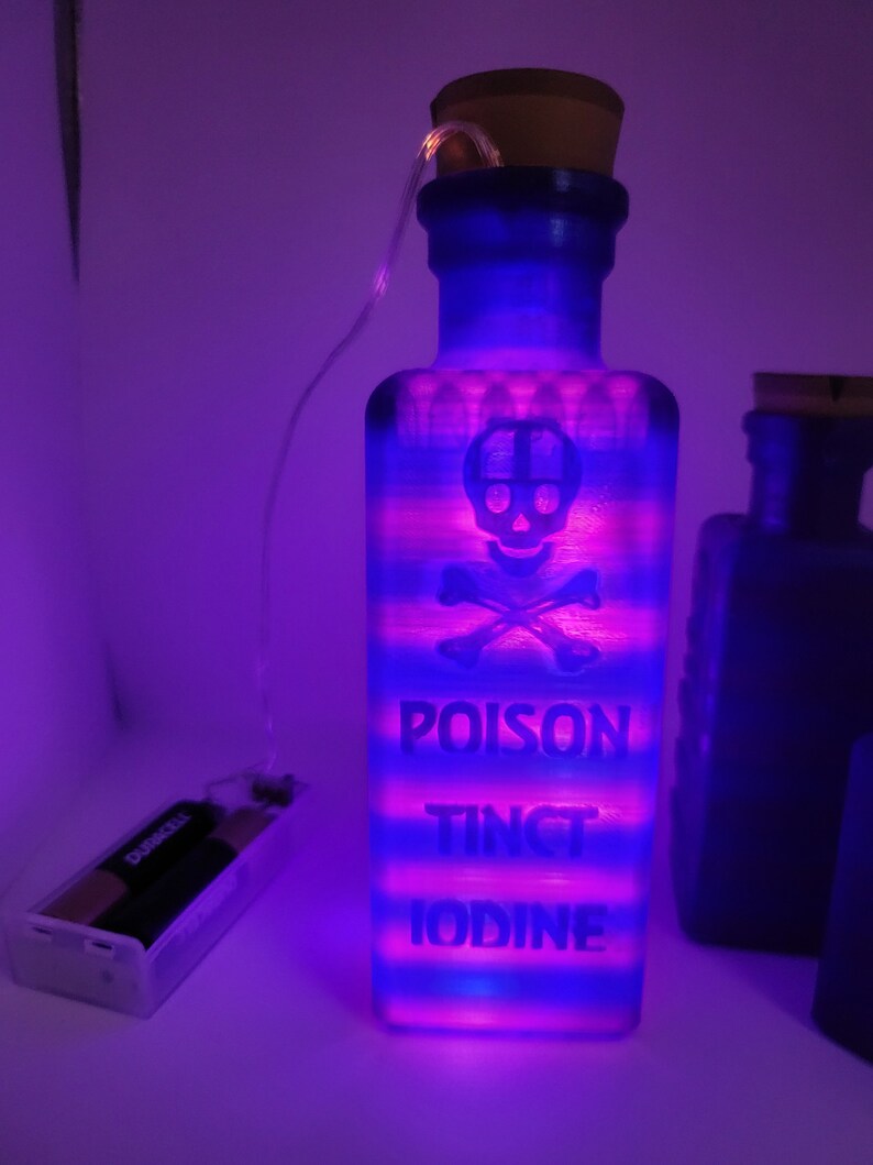 May include: A blue glass bottle with a cork stopper and a skull and crossbones design. The bottle is lit from within with pink and blue lights and has the words "POISON TINCT IODINE" printed on it.