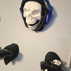 May include: A white skull wearing a black hood and blue headphones with glowing lights. Below, two black hooded figures hold black game controllers with white skeletal hands. The wall art has a gaming theme.
