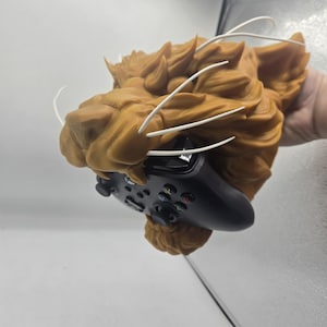 May include: A tan-colored lion head sculpture with white whiskers, holding a black Xbox controller. The sculpture has detailed fur and is designed to hold the controller in its mouth. The controller has multiple buttons and joysticks.