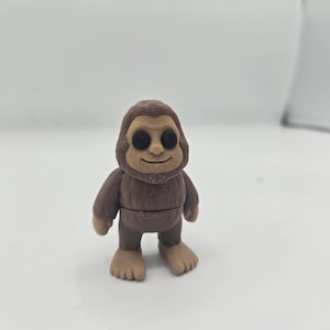 May include: A brown figurine of a cartoon Bigfoot with a smiling face and large black eyes. The figure has a brown hooded sweatshirt and tan feet. The figurine is standing on a white surface.