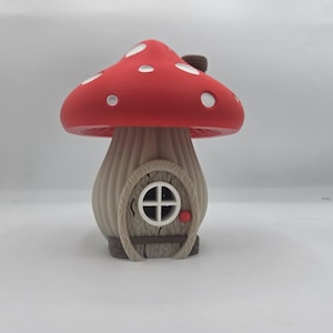 May include: A mushroom-shaped house figurine featuring a bright red cap with white spots and a light brown, textured stalk. The stalk includes a central arched door with a small white circular window and a tiny red knob, resembling a dwelling.